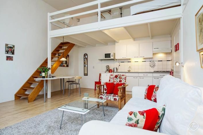 lit mezzanine blanc au dessus d une cuisine kitchenette blanche ouverte sur salon avec canapé blanc et table en verre sur tapis gris sur parquet bois clair