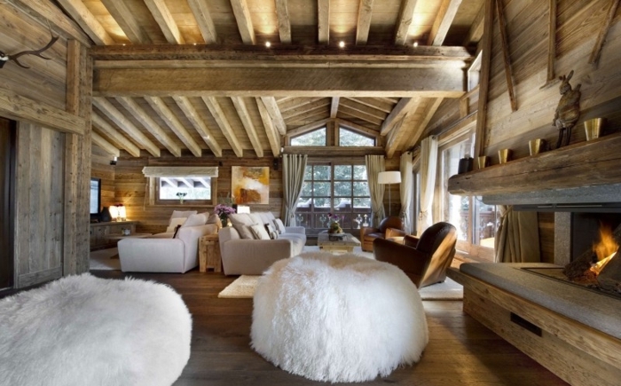  canapé cocooning, plafond avec poutres en bois, grand pouf blanc, cheminée allumée, grandes fenêtres