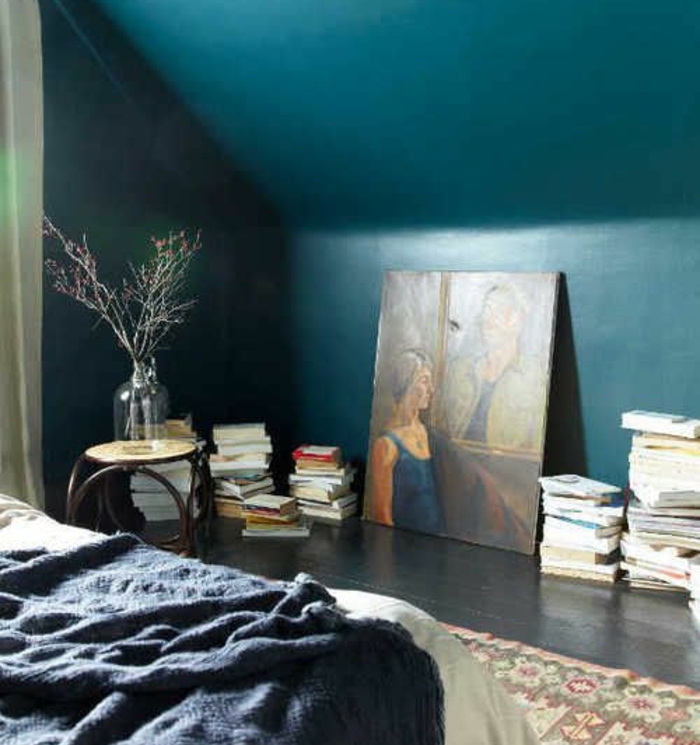 chambre mansardée, couleur mur bleu paon, parquet en bois gris anthracite, piles de livres, tableau peinture, tapis oriental, couverture de lit gris anthracite