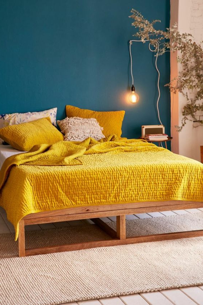 mur bleu canard, lit avec plaid jaune moutarde, ampoule et petit chevet 