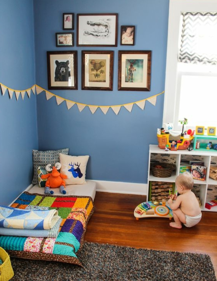 idée déco chambre enfant, mur couleur bleue, deco murale, lit bébé montessori au sol, tapis gris, meuble de rangement bas, couverture de lit multicolore, jouets, coussins