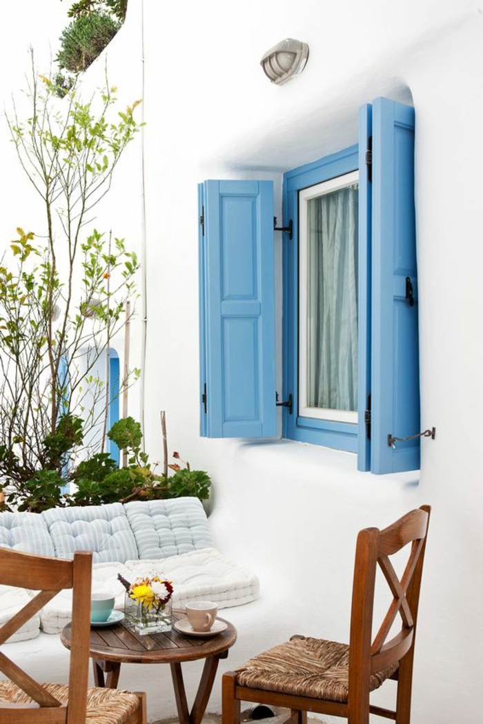 bleu grec sur les volets, salon de jardin en bois, fleurs vertes, façade blanche