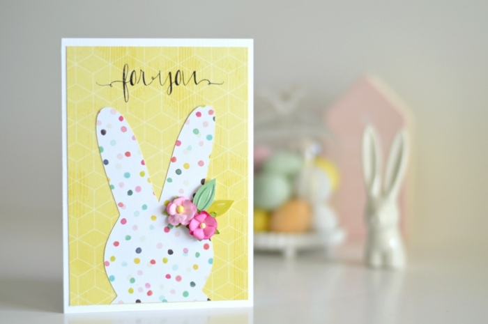 carte de paques, silhouette de lapin de paques blanc à points multicolores, fleurs rose, fond papier jaune