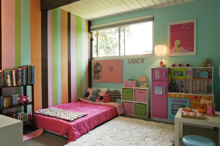 mur couleur verte, et mur d accent à rayures multicolores, lit au sol, couverture de lit rose, tapis blanc cassé, bibliothèque livres, petite cuisine enfant, rangements, chambre montessori fille