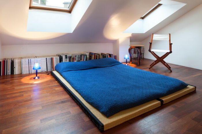 lit à même le sol, parquet en bois marron, linge de lit bleu, rangement livres sur le sol, chaise pliable, peinture mur blanc, déco chambre sous pente