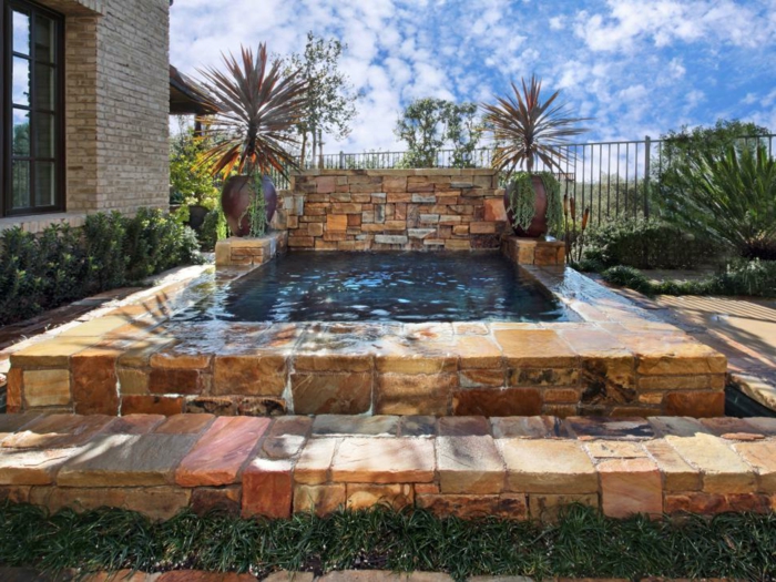 piscine surélevée en pierre, gazon, plantes vertes, palmier, maison en briques