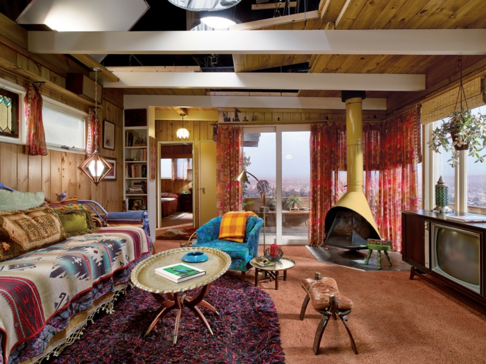 chambre hippie, murs en bois, tapis ethnique, cheminée moderne, fauteuil bleu foncé, grande fenêtre