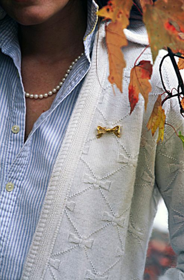 collier de perles porté avec une veste blanche aux motifs noeuds repris sur la broche en métal jaune 