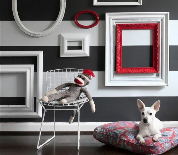 mur à rayures décorée de cadres vides ovales et rectangulaires en blanc et rouge, petit chien blanc sur un coussin, chaise en métal, jouet singe, habiller un mur