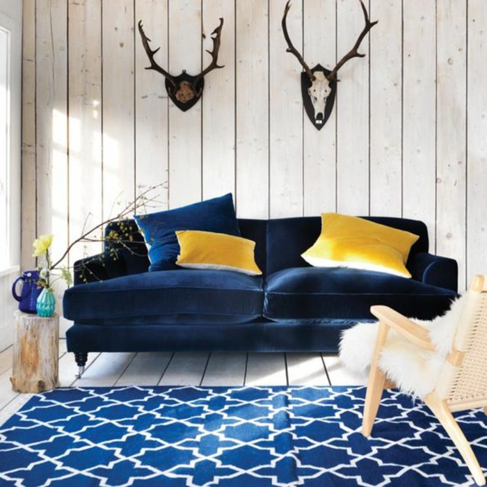 deco salon scandinave, canapé et tapis bleu marine, lambris bois, trophées de chasse, chaise scandinave avec fourrure, bûche de bois décoratif, coussins jaunes