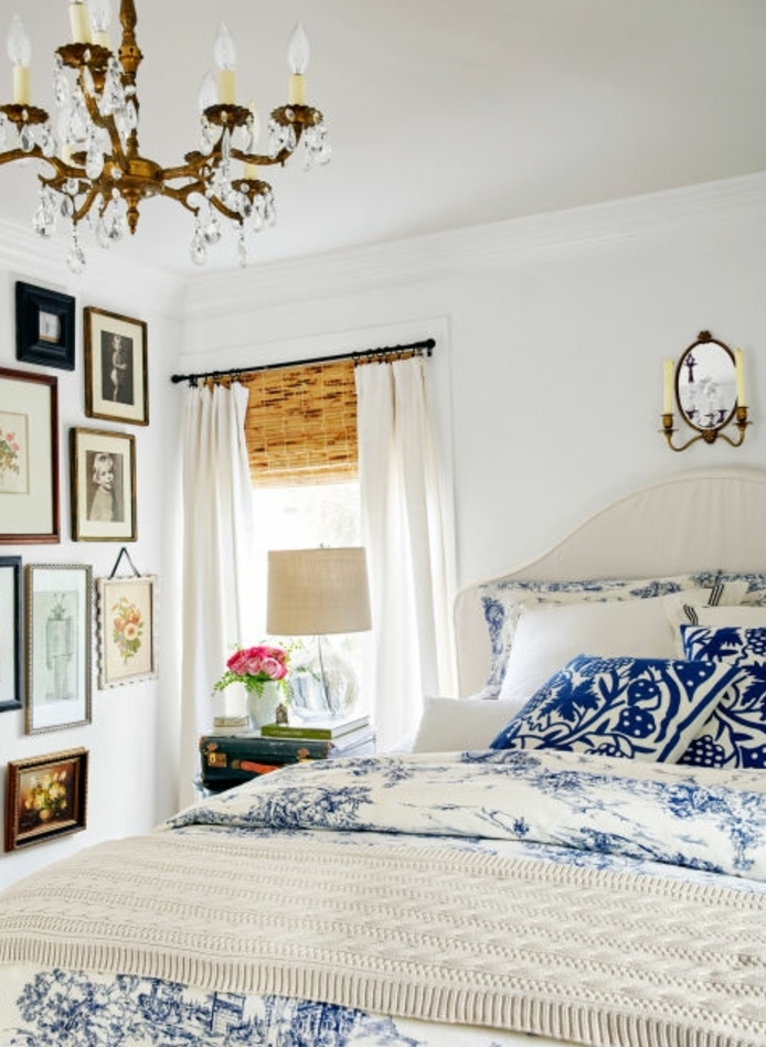 chambre a coucher deco campagne, linge delit blanc à motifs bleus, deco murale de photo et dessins, lustre baroque, rideaux blancs légers, table de nuit malle vintage