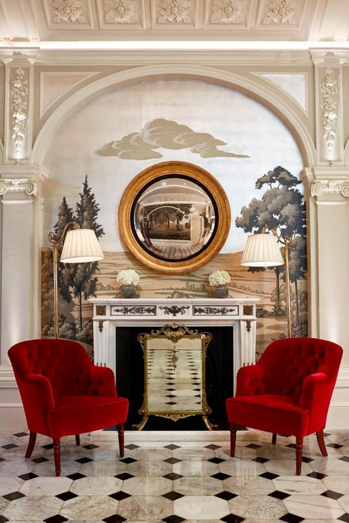 miroir vintage oeil de sorcière sur un mur peint avec un tableau de nature foret et champs et deux fauteuils rouges classiques des deux cotés de la cheminée