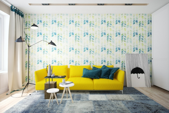 salon scandinave, mur habillé de papier peint à motifs floraux, bleu, jaune et vert, canapé jaune, tapis gris et bleu, tables scandinaves, parquet clair, lampe design