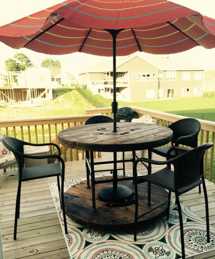amenagement terrasse en teck, touret bois table vintage, un parasol qui passe par le centre, chaises noires, tapis à motifs orientaux