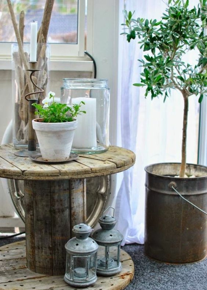 touret bois decoration, table bois brut, lanternes vintage, pot de fleur en terre cuite blanchi, bocal en verre avec bougie à l interieur, arbre planté dans un seau en metal