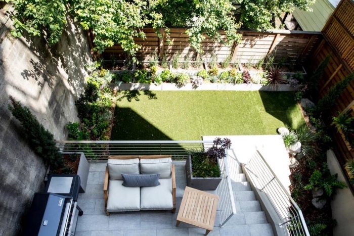 une terrasse en béton avec cuisine d été, barbecue fauteuil en bois et plants dans des pots en beton, gazon avec bordure d arbustes