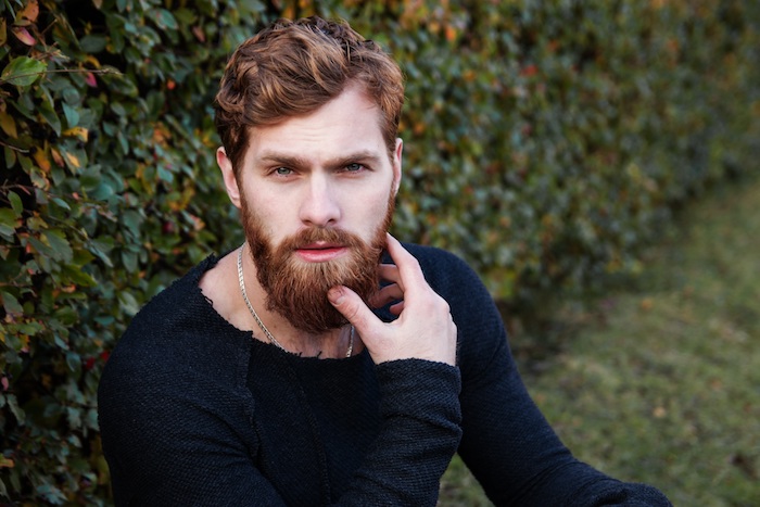 coupe-de-cheveux-homme-tendance-barbe-longue-couleur-de-cheveux-brun-châtain-clair homme brun, blouse noire aux manches longues, collier en argent homme, cheveux ondulés et barbe longue en cuivre