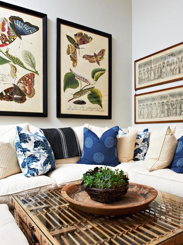 tissu tropical, salon aux murs blancs, coussins décoratifs bleus, canapé d'angle beige, plant verte, peinture papillon aux cadres noirs