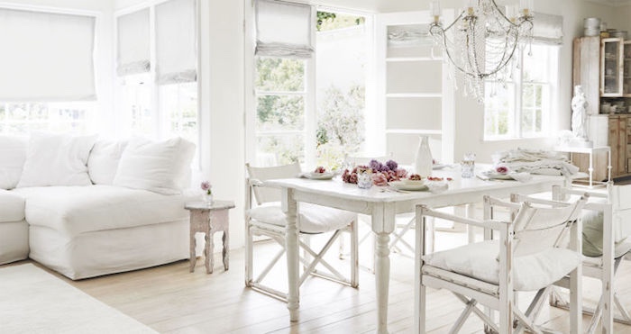 intérieur tout en blanc, parquet clair, canapé blanc, table et chaises blanches usées, lustre baroque, decoration de fleurs colorées sur al table