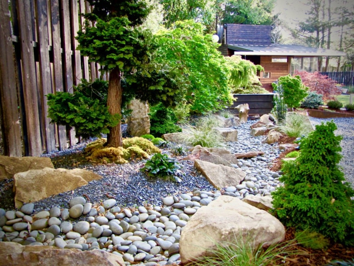 jardin zen japonais avec une riviere en galets, entourée de grosses pierres, arbres et arbustes verts, clôture en bois, maison dans les bois, chalet design