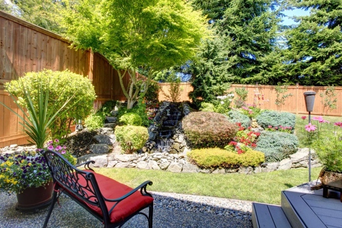 idee deco jardin avec gazon, cascade d eau dans une rockaille fleurie, d arbustes et d arbres, un banc en metal avec coussin d assise rouge