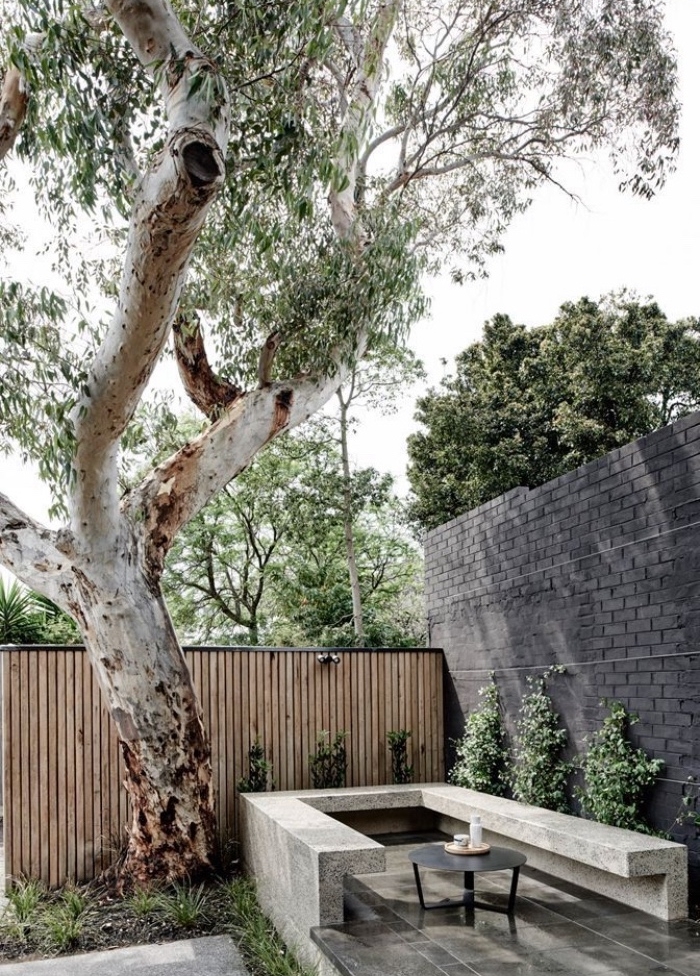 idee amenagement jardin sec, espace détente, banc en béton et petit table basse metallique noire, arbre à couronne verte, clôture en bois, arbustes 