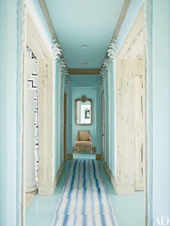 couloir couleur vert pastel et blanc, tapis à rayures blancs et bleus, un fauteuil beige au fond et miroir baroque