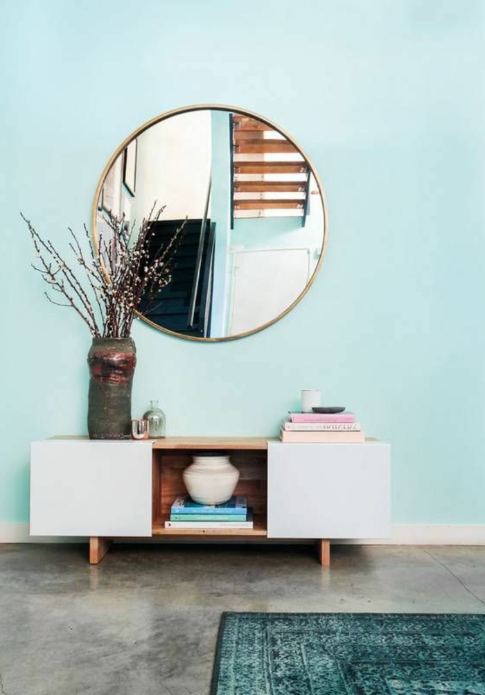 peinture vert d eau sur le mur, grand miroir rond, meuble de rangement, revêtement aspect béton, tapis bleu canard et branches dans un vase