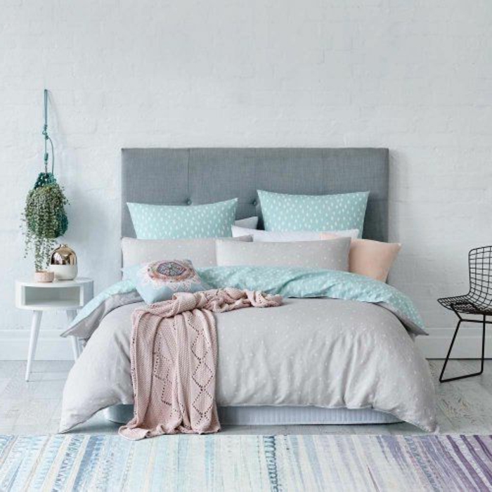 lit gris, couvert de linge de lit couleur vert pastel aqua, gris et rose, tapis à rayures couleurs usées, mur en briques blanches, tête de lit grise, plante suspendue