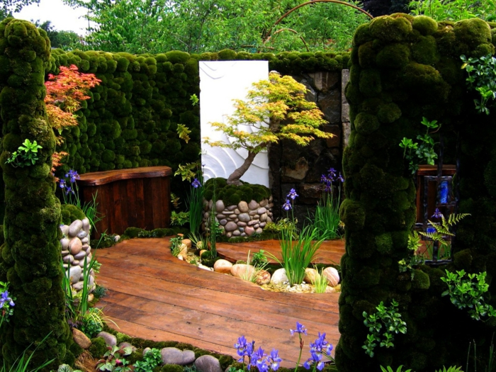 un coin détente cloisonné d un mur de mousse verte, sol et banc en bois, arbre et fleurs fraiches, pauysage paradisiaque