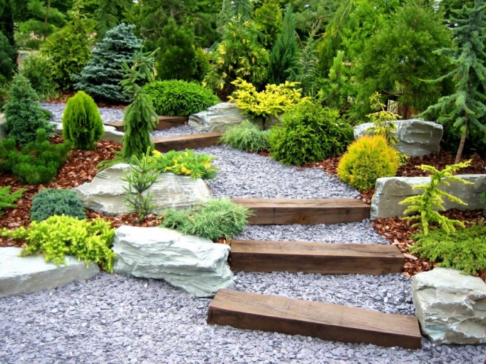 un escalier en blocks de bois, submergés de gravier, chemin entouré de pierres, arbustes et arbres, végétation verte