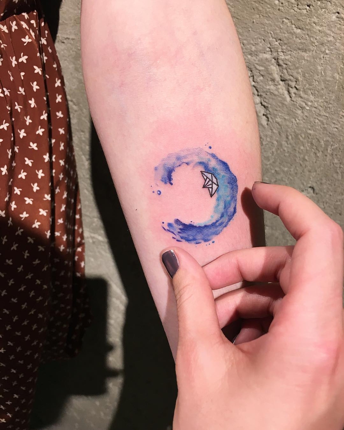 un tatouage femme bras de style aquarelle représentant un petit bateau en papier et une vague