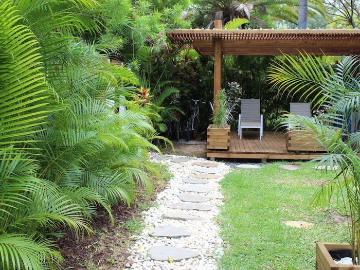 amenagement terrasse exterieure, pergola bois et revêtement composite, chaise longues bleu et gris, gazon vert et mur de palmiers, chemin de pierres