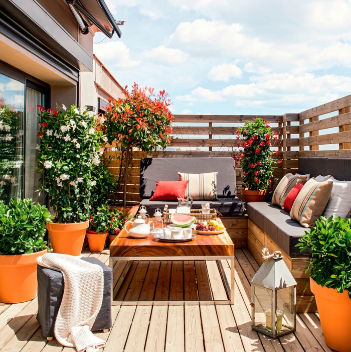 aménager une terrasse avec un canapé d angle en bois et coussins d assise marron, table en bois, plantes dans des pots de fleurs, arbres, revêtement en bois, lanterne grise