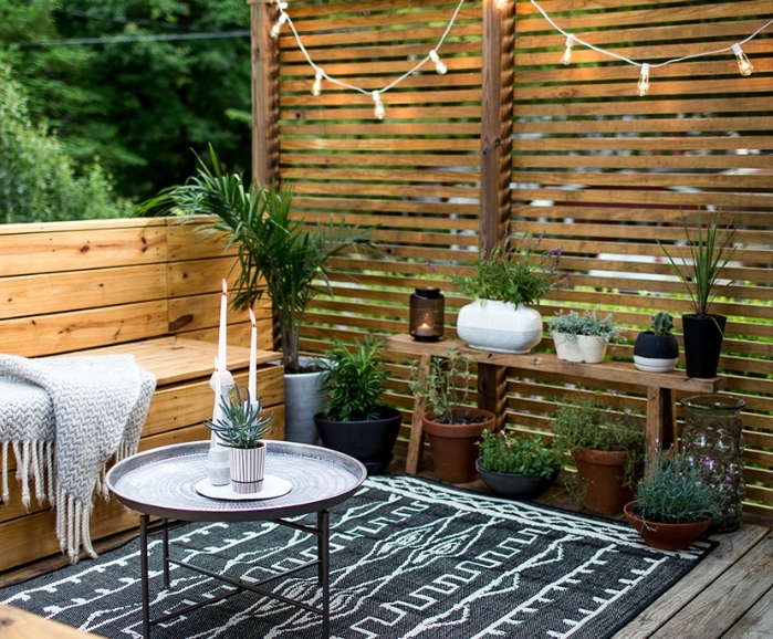 deco terrasse en bois, canapé et brise vue en bois, tapis noir à motifs géométriques blancs, table basse design, plaid gris, plantes vertes, guirlande lumineuse