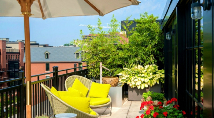 deco terrasse sur un petit balcon avec revêtement en béton, chaises en rotin avec coussins jaunes, parasol, bambou, plantes et fleurs, petite fontaine