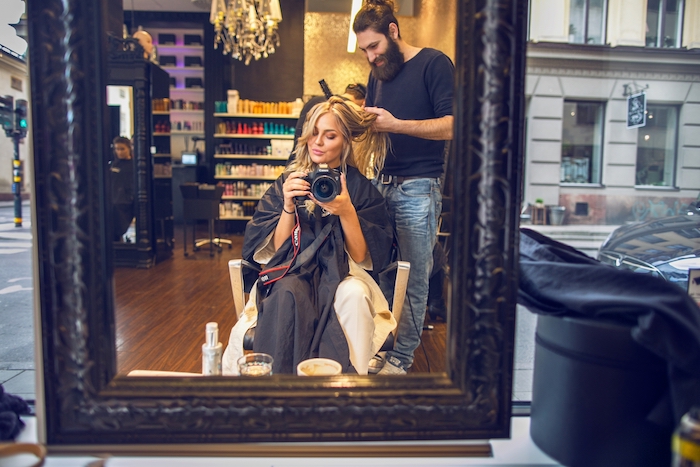 balayage-blond-appareil-photo-visite-coiffureur-salon-de-beauté-cheveux-longs-pantalon-blanc meche blonde, femme devant grand miroir, soins de beauté cheveux, visite salon coiffeur, lustre en verre et cristaux