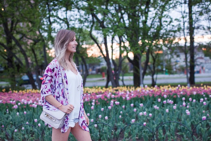 balayage-ombre-blonde-jardin-de-tulipes-promenade-parc-femme-shorts-en-denim-débardeur-blanc-collier-en-or coloration cheveux, sac à main blanc avec poignée longue, shorts en denim et gilet violet, coupe de cheveux mi longs