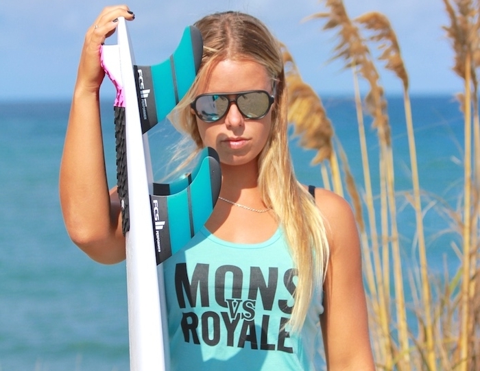 blond-doré-manucure-noire-bague-métalique-lunettes-de-soleil-effet-miroir-cheuvex-longs californien cheveux, débardeur bleu avec lettres noires, ongles courts peints en noir, planche de surf blanche