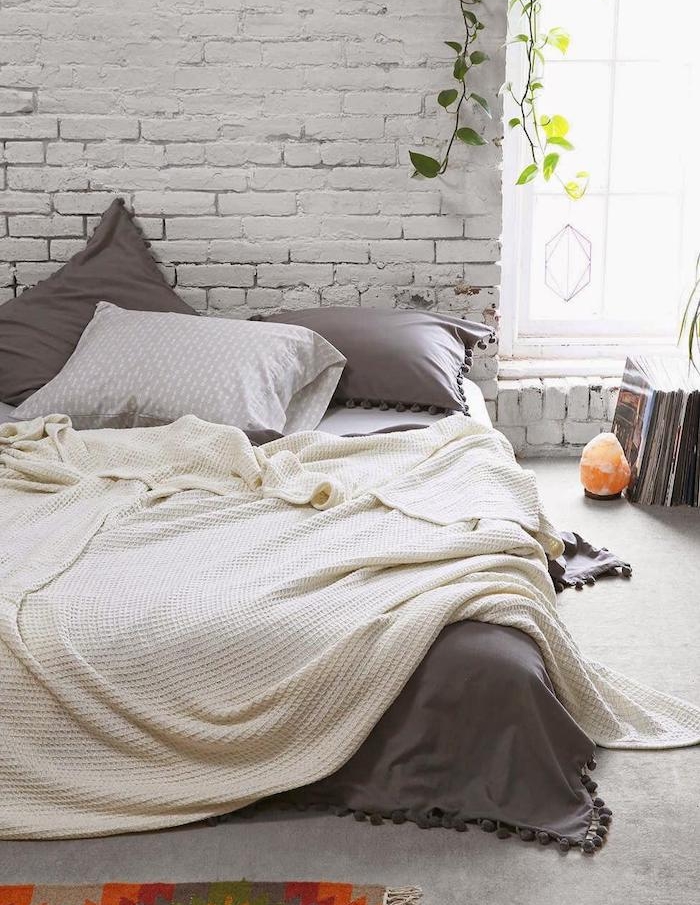 modele de chambre cocooning, mur en briques, lit matelas avec linge de lit blanc et gris, plantes vertes et magazines par terre