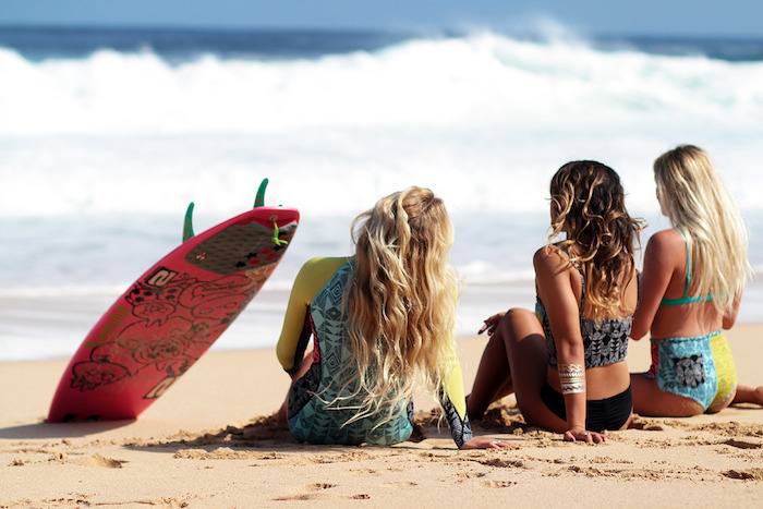 balayage sur cheveux chatain, amies sur la plage, planche de surf en rouge avec dessins noirs, maillots de bains deux pièces en jaune et bleu