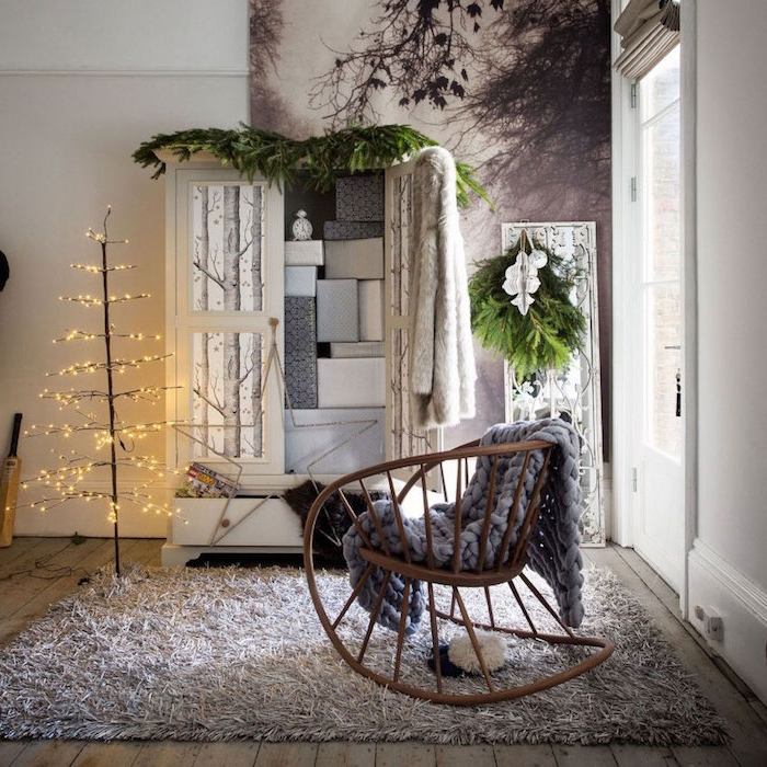tapis cocooning dans une habitation hygge, chaise à bascule en bois, plaid gris à grosses mailles, armoire blanche, arbre noel avec guirlande lumineuse, panneau decoratif motif foret nordique