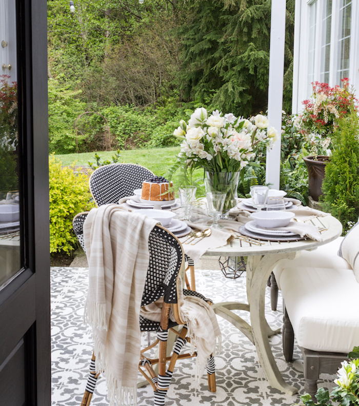 une petite salle à manger sur une terrasse zen, revêtement carrelage mosaique blanc et gris, table blanche, canapé bois avec coussin d assise blanc, chaises en bois, tissu blanc et noir