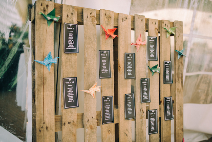 listes invités noires, lettres blancs sur une palette bois rustique et papier origami coloré, idée plan de table mariage original