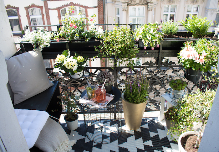petit balcon, amenagement terrasse minuscule, assise en bois, coussins et couverture gris, tapis noir et blanc, pots de fleurs, plantes vertes, table en poubelle retournée