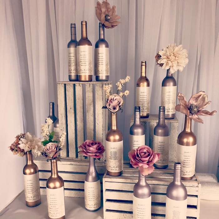 plan de table mariage avec des bouteilles de vin argentée et dorées disposées autour de caisses de bois blanchis, aspect usé et petites fleurs dedans