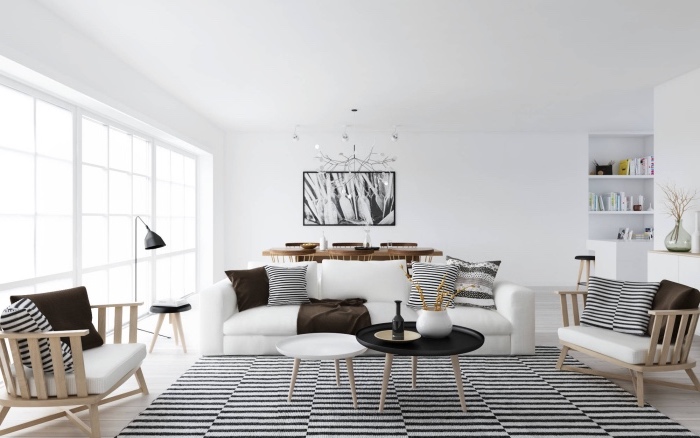 salon moderne dans une maison scandinave, tapis noir et blanc, canapé blanc, fauteuils nbois avec coussins d assise blancs, coussins decoratifs noir et blanc et marron, chaises et table en bois et decoration murale peinture graphique