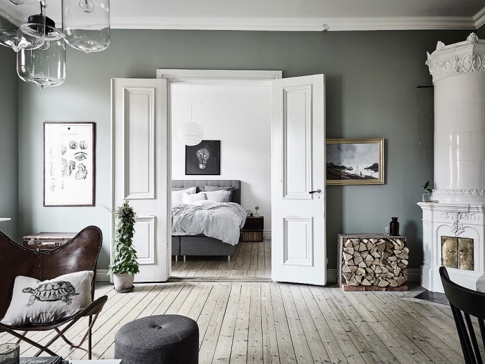 chambre à coucher ouverte sur salon, parquet gris clair, lit gris avec linge de lit blanc cassé, chaise cuir, cheminée blanche, intérieur rustique