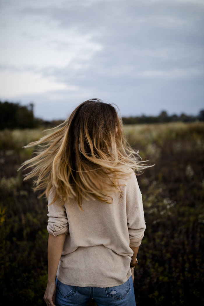 ombré-hair-blond-blouse-femme-beige-paire-de-jeans-claires-femme-dans-la-nature-champs balayage sur cheveux chatain, blouse beige et pantalon en denim modèle femme, coiffure avec racines foncées