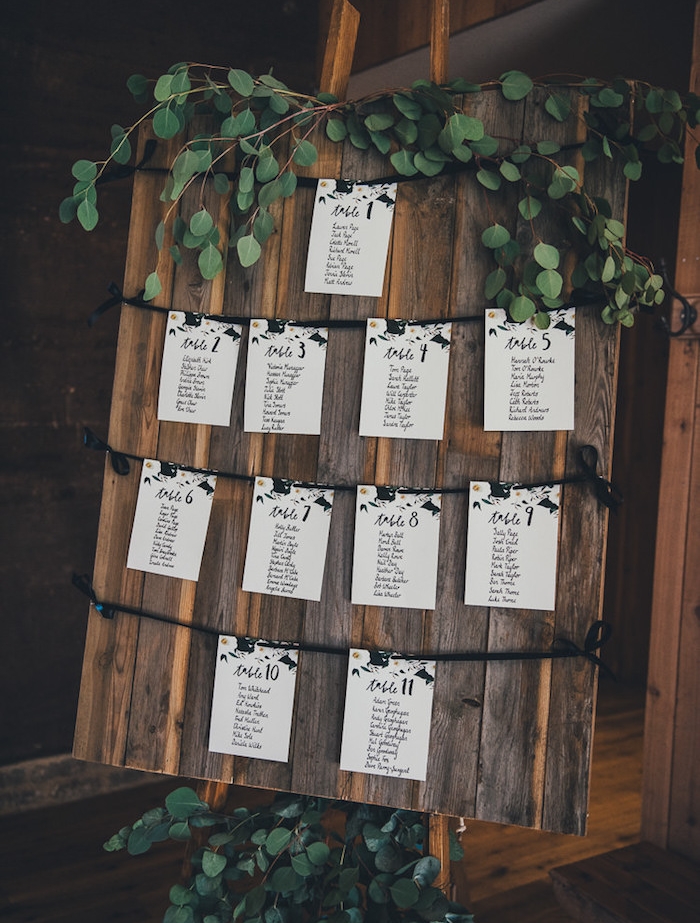 plan de table pour mariage en planches de bois vintage marron et petites listes invités blanches, écriture calligraphie en lettres noires, guirlande vert